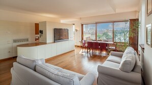 Eine moderne Küche mit einer zentralen Kochinsel, einem Essbereich mit rotem Tisch und Stühlen sowie einem großen Fenster mit Blick auf die Stadt.