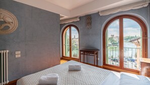 Ein Schlafzimmer mit einem großen Bett, zwei Fenstern, Blick auf eine Stadtlandschaft und einer Heizung.