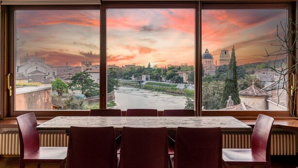 A dining area with a large window overlooking a river and historic buildings during sunset. - Ponte Pietra Residence (Verona)