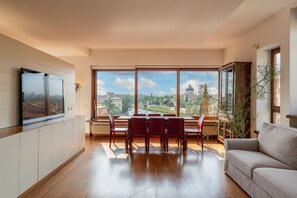 A spacious living room with a dining area, large windows, a flat-screen TV, and a view of a cityscape.