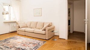 A living room with a beige sofa, a small round table, a colorful rug, a radiator, and a door leading to another room.