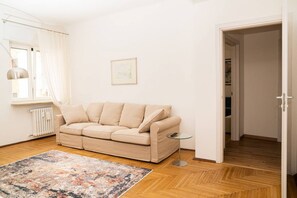 A living room with a beige sofa, a small round table, a colorful rug, a radiator, and a door leading to another room.