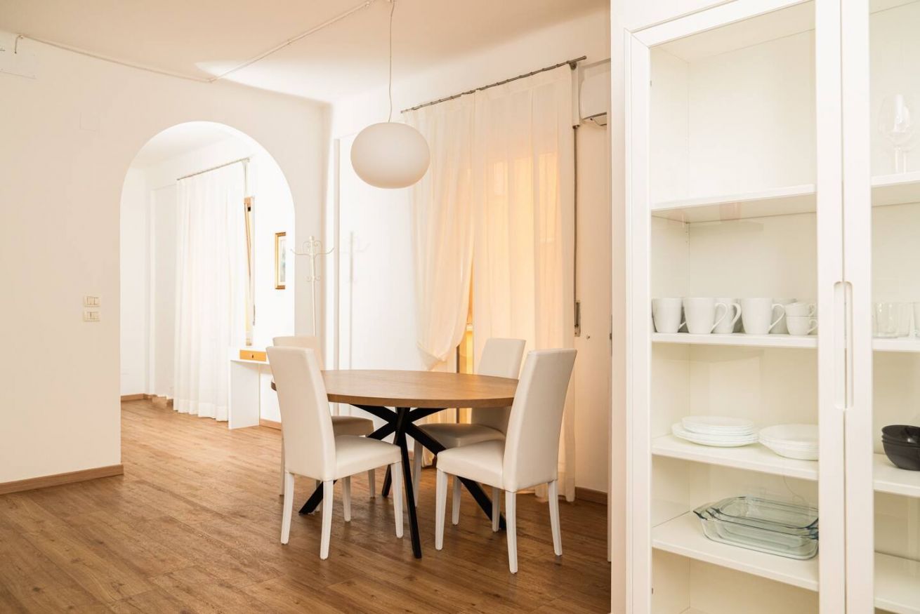 A dining area with a round table, four chairs, and a glass-top table. There is a white cabinet with shelves on the right.