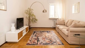A modern living room with a beige sofa, a flat-screen TV on a white console, a large abstract rug, a potted plant, and a floor lamp. - Serego Residence - Elegant residence overlooking Porta Leoni (Verona)