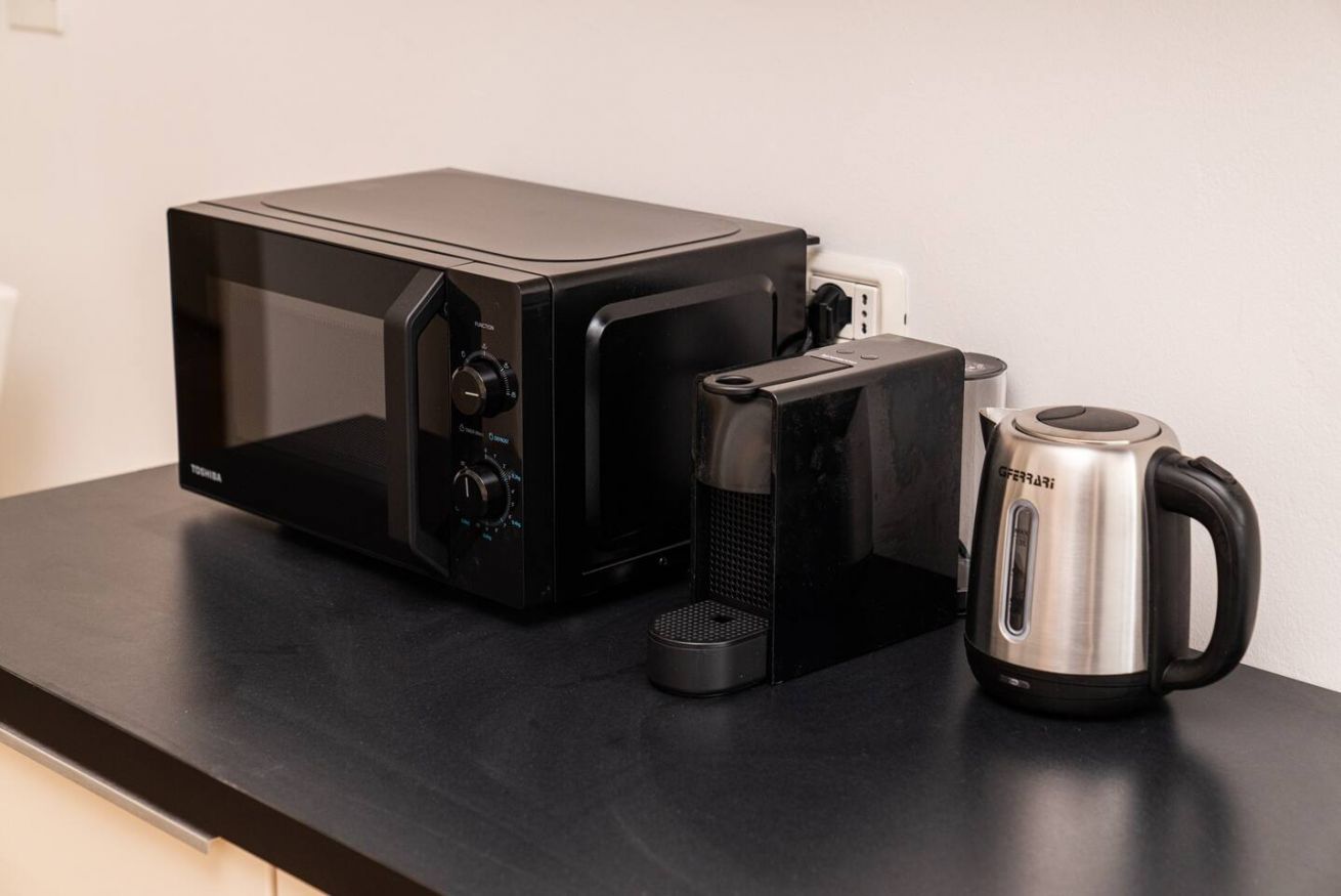 Et køkken med en sort mikrobølgeovn, kaffemaskine og elkedel på en sort bordplade.