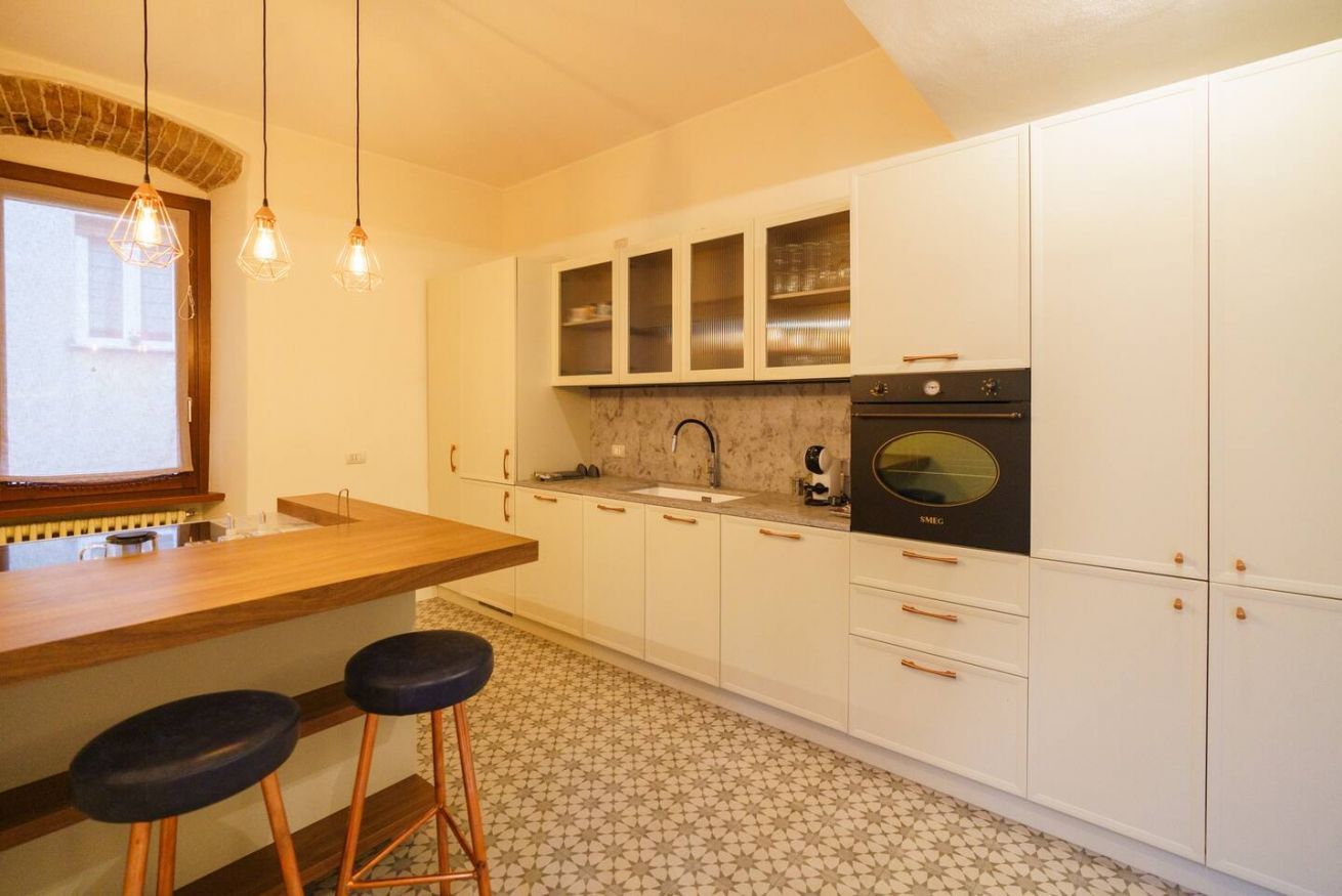A compact kitchen with white cabinets, a wooden countertop, a built-in oven, and two stools.