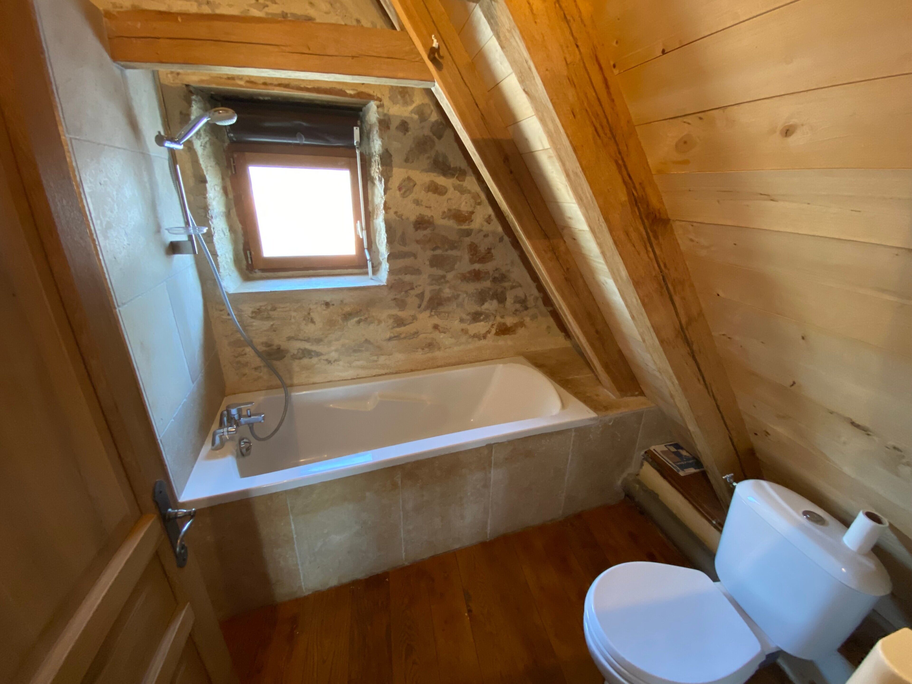 Une petite salle de bain avec un toit en pente, une baignoire, des toilettes et une fenêtre située au-dessus de la baignoire.