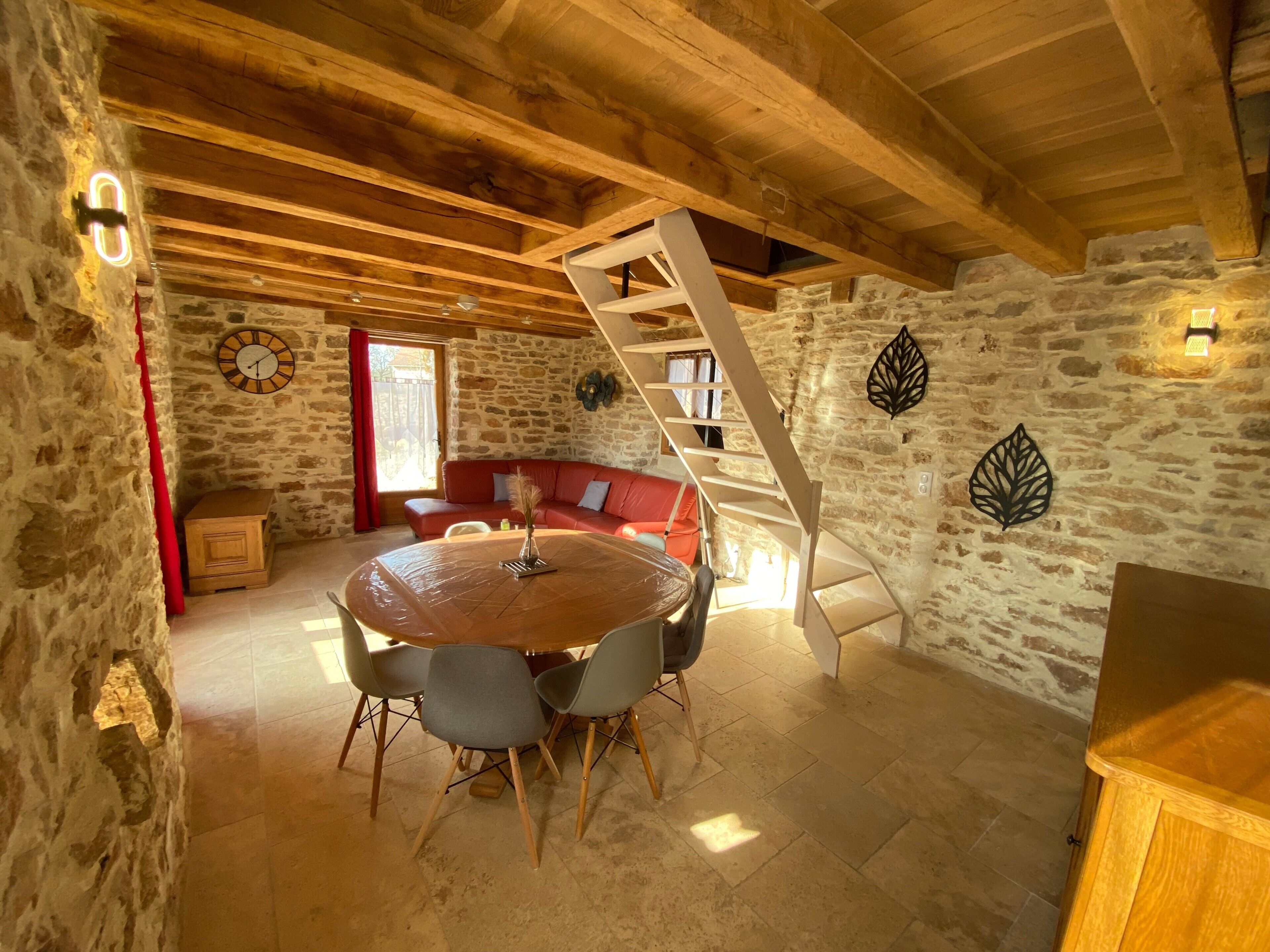 A rustic room with a stone wall, wooden ceiling beams, a round wooden table with chairs, a red sofa, and a wooden ladder.