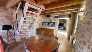 Een rustieke kamer met een houten tafel, stoelen, een wenteltrap, stenen muren en een open haard.