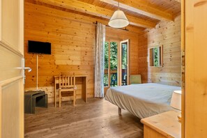 A wooden cabin room with a bed, a chair, a desk, a television, and a window.