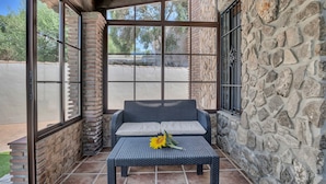 A covered patio area with a stone wall, a wicker sofa with cushions, and a small table with a sunflower.