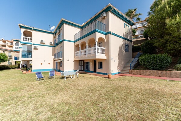 A multi-story building with balconies, a lawn with chairs, and a staircase leading to a lower level.