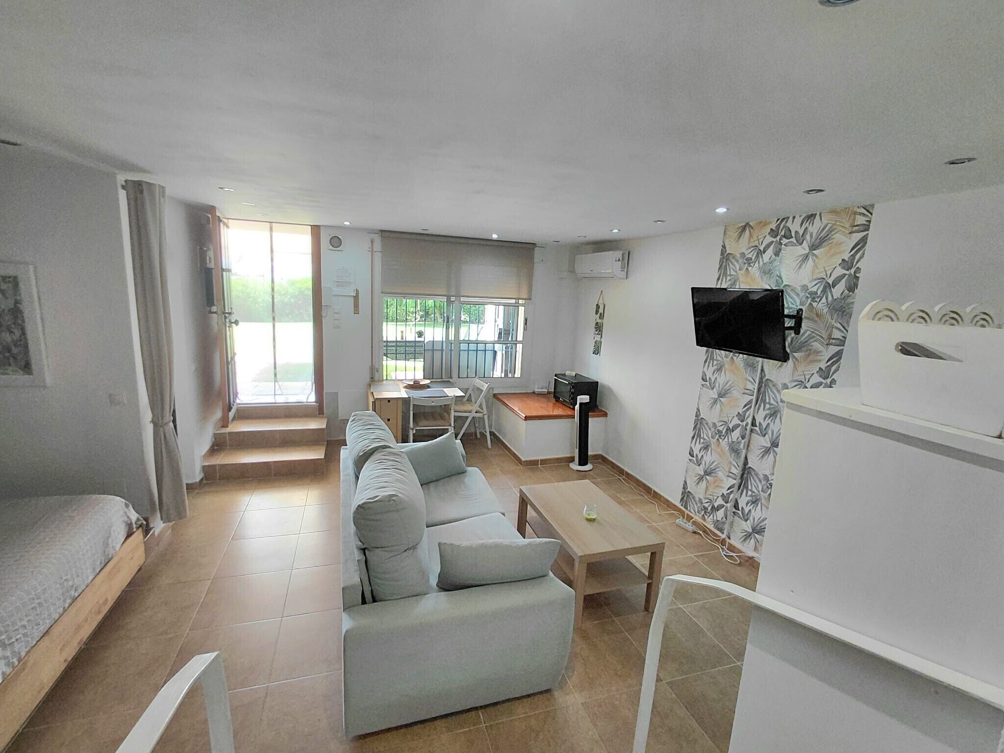 A modern hotel room with a sofa, a small table, a TV mounted on the wall, and a balcony with a view.
