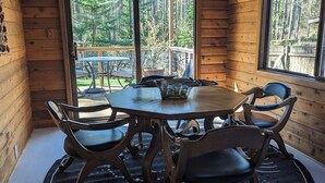 Un interior de madera con una mesa redonda y sillas, ventanas amplias y vistas a un bosque.