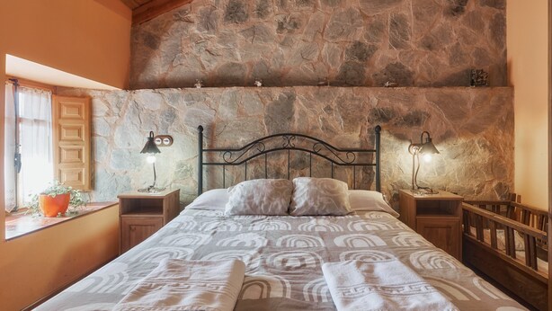 Une chambre avec un mur en pierre, un lit recouvert d’une couvre-lit à motifs, deux lampes de chevet et un berceau en bois.