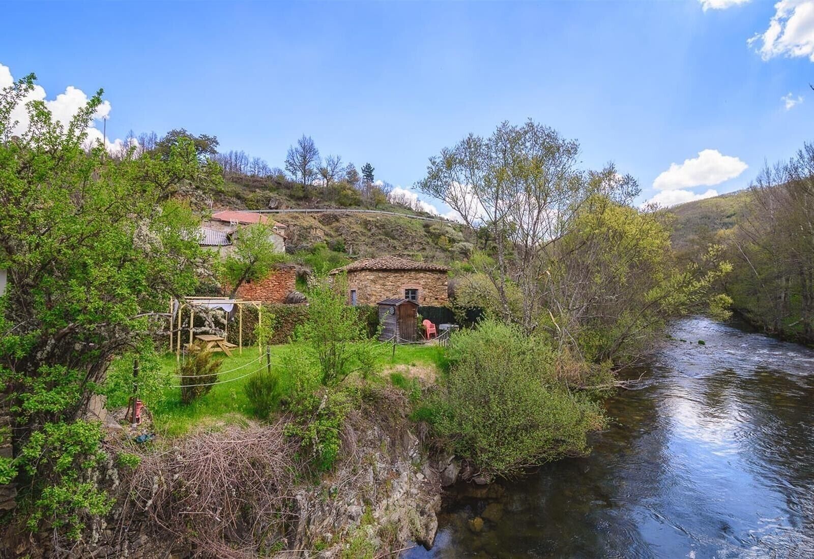 Un cadre rural au bord de la rivière, avec des bâtiments en pierre, une structure en bois et une végétation luxuriante.
