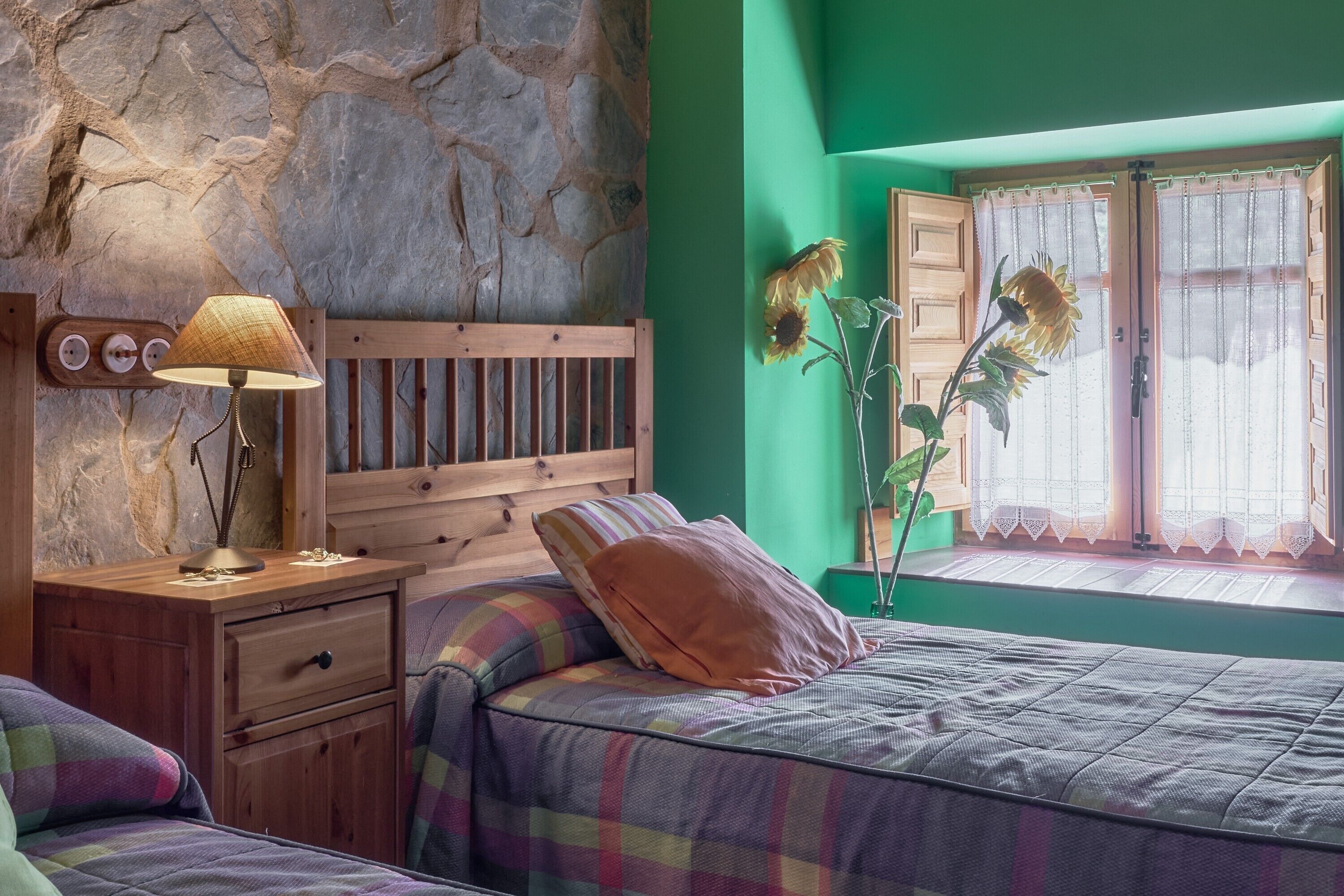 Een slaapkamer met een stenen muur, een houten hoofdbord, een nachtkastje met een lamp, een bed met ruitdessin beddengoed en een raam met zonnebloemen.