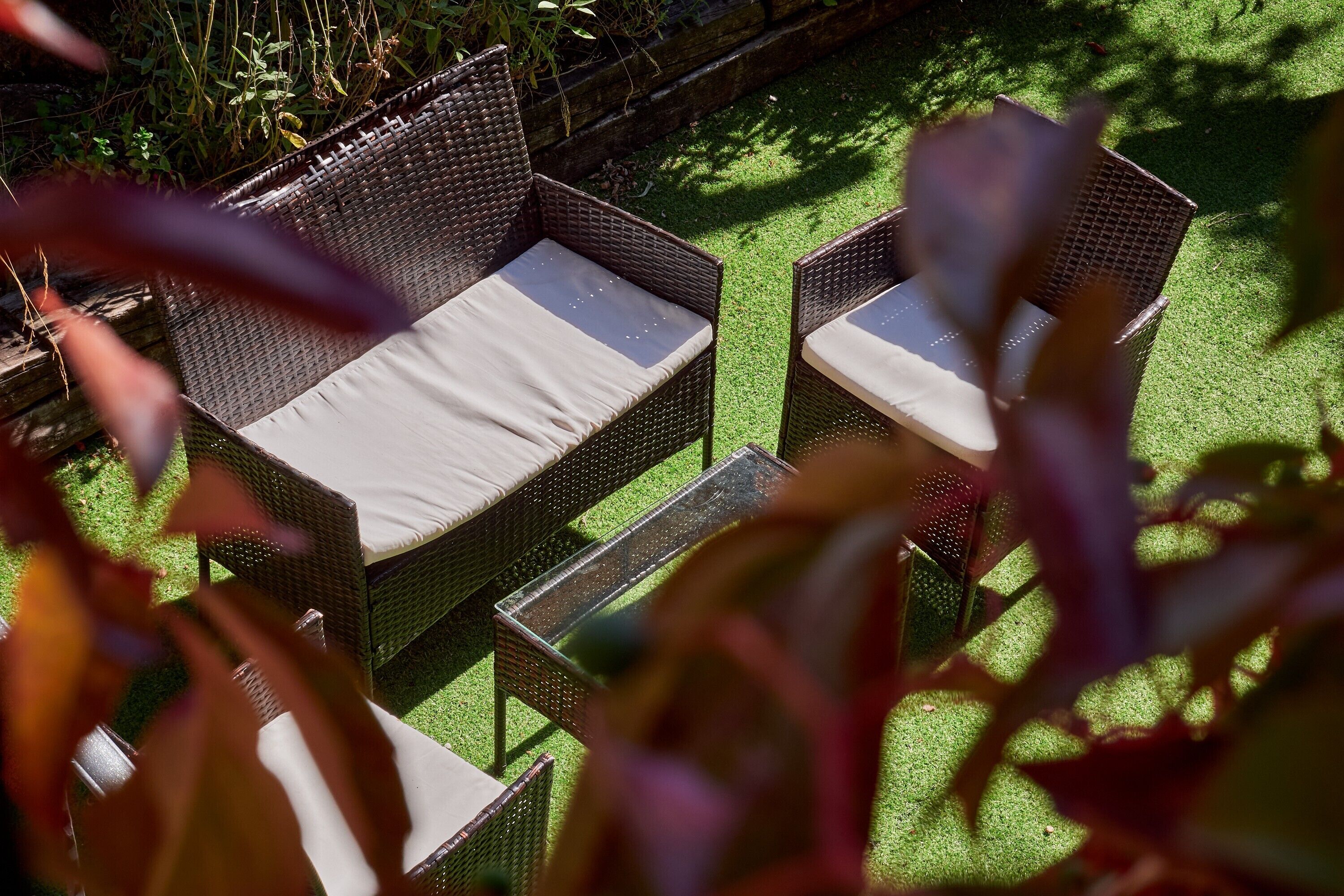 Un espace de détente extérieur aménagé avec des meubles en osier, des coussins, une table en verre et une pelouse verdoyante.