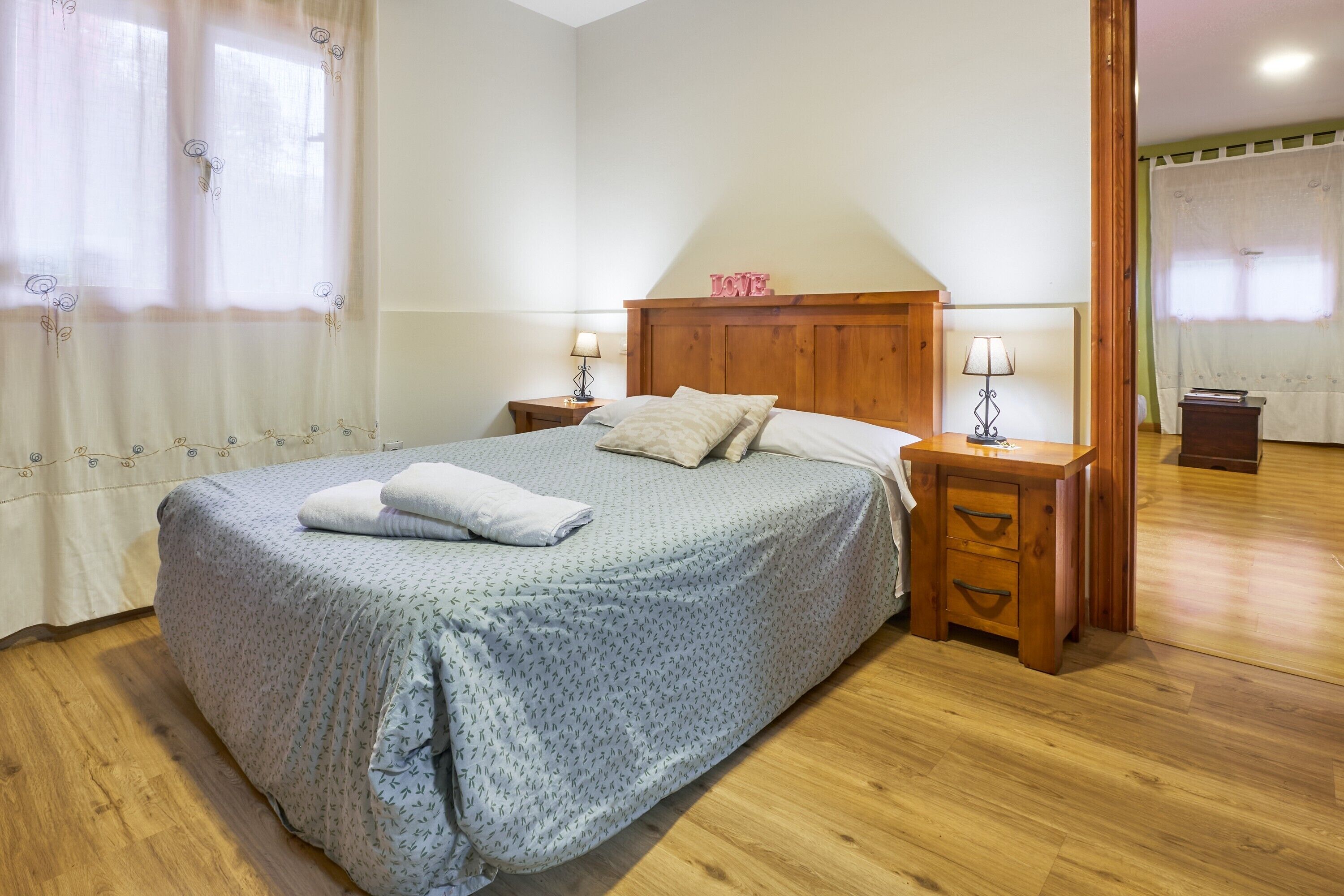 Een net opgemaakt bed met een houten hoofdbord, twee nachtkastjes met lampen, en een klein houten bijzettafeltje.