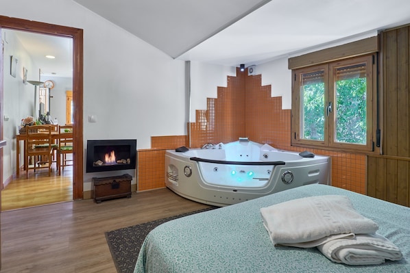 A bedroom with a large bed, a fireplace, a jacuzzi, and a window with a view.