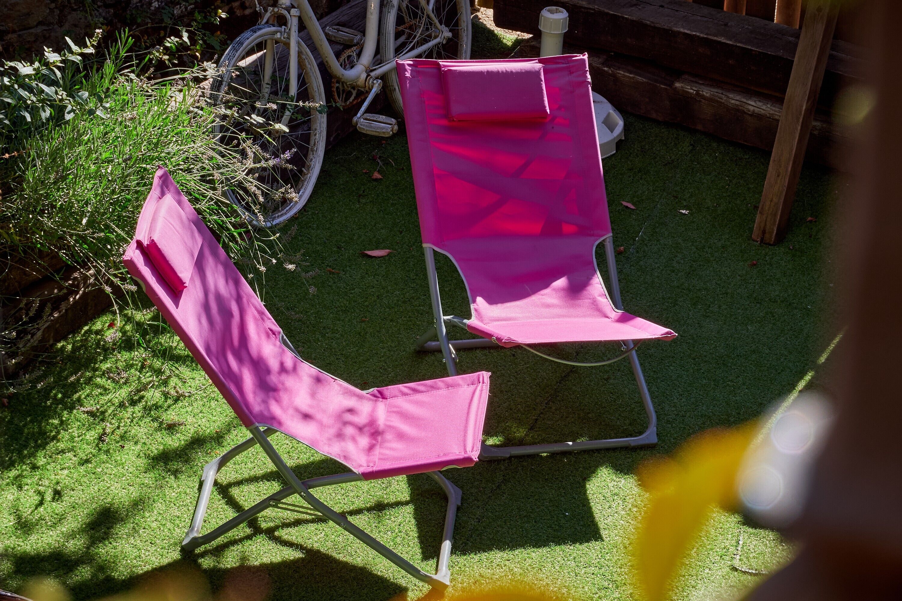 Two pink lawn chairs on artificial grass with a bicycle and greenery in the background.