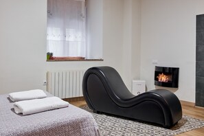 A modern room with a massage chair, a bed with towels, a fireplace, and a window with sheer curtains.