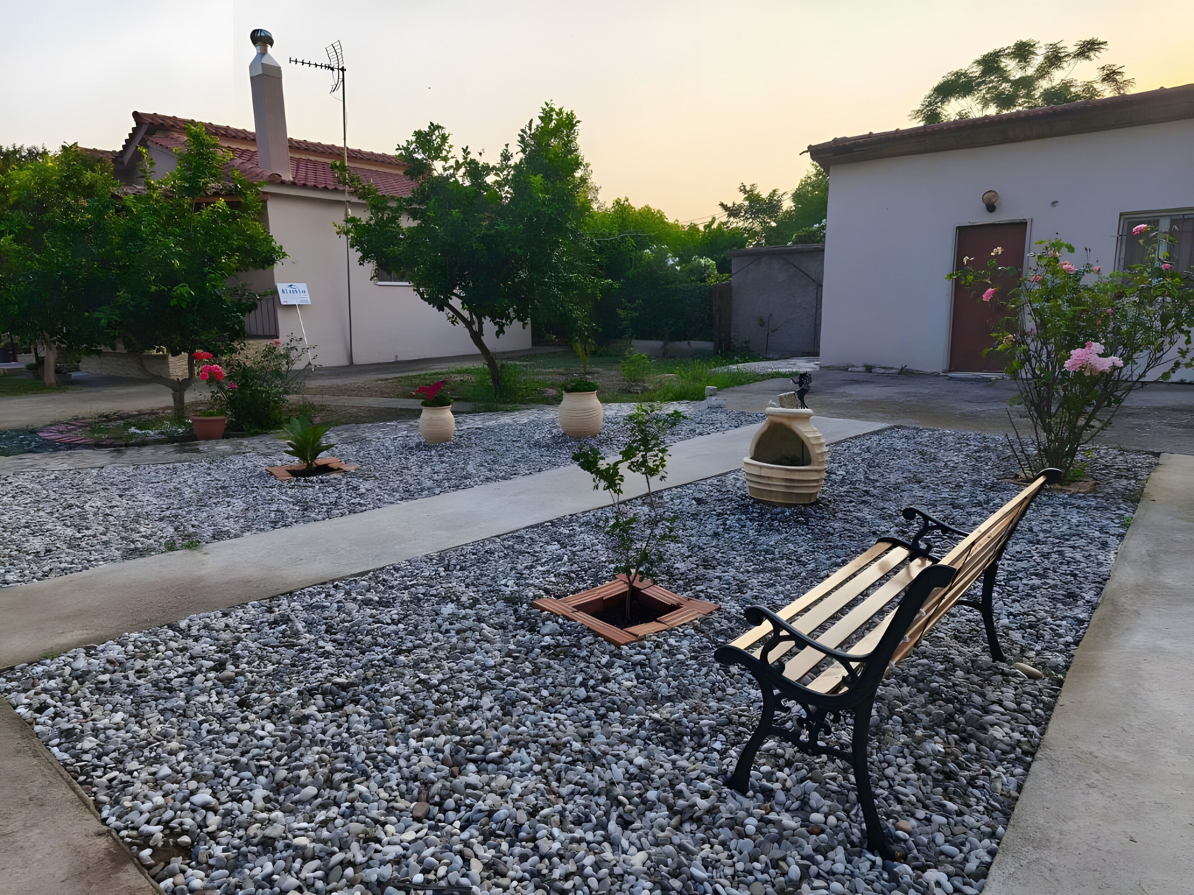 A courtyard with a gravel path, a wooden bench, potted plants, and a building with a tiled roof.