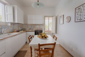 Une cuisine moderne avec des meubles blancs, une table à manger avec des chaises, un panier de fruits et une fenêtre donnant sur un beau paysage.