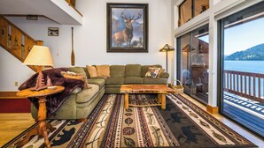 A cozy living room with a green sofa, a wooden coffee table, and a patterned rug. A large framed deer painting adorns the wall, and a window offers a view of a deck and water.