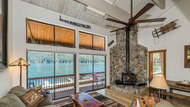 A cozy living room with a stone fireplace, large windows overlooking a lake, and a ceiling fan.