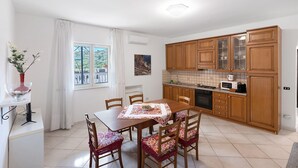 A dining area with a table and chairs, a kitchen with wooden cabinets, and a window with curtains. - Apartment 'Appartamento Capri' with Sea View, Wi-Fi and Air Conditioning (Massa Lubrense)