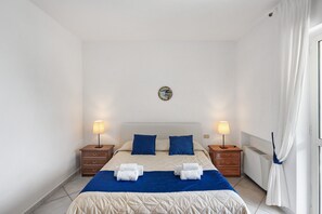 A double bed with a blue and white duvet, two bedside tables with lamps, a wall clock, and a window with curtains.