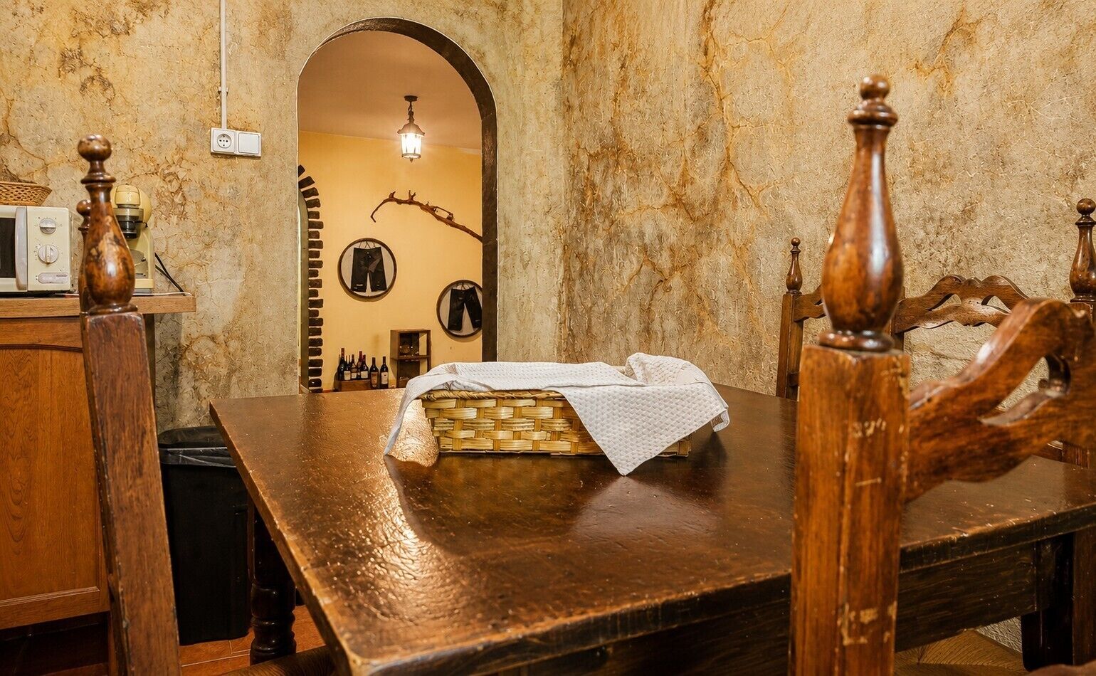 Een houten eettafel met een gevlochten mand en een witte tafelkleed, een houten stoel met een gebeeldhouwd rugleuning, en een muur met een ronde spiegel en lijstwerken.