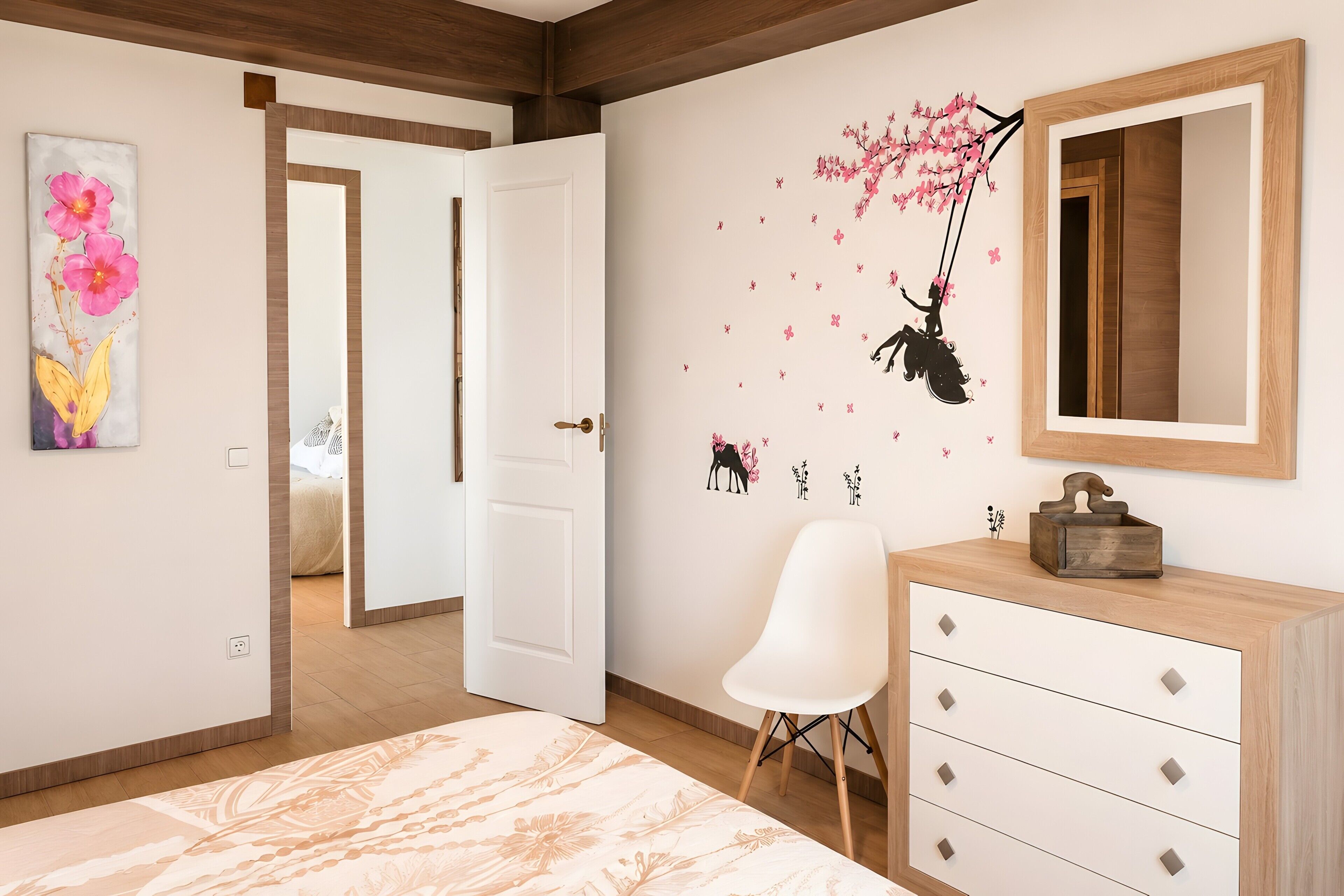 A bedroom with a bed, a dresser, a chair, a door, and a wall with a floral painting and a wall decal.