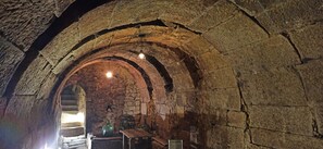 Ein gewölbter Innenraum aus Stein mit hängenden Lampen, einem Holztisch und einer grünen Flasche.