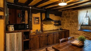 A rustic kitchen with wooden cabinets, a stone wall, a window with curtains, and a dining table with chairs.