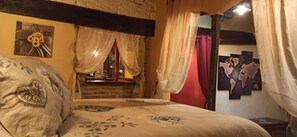 Een slaapkamer met een bed, een raam met gordijnen, een aan de muur hangend schilderij en een klein tafeltje met een lamp.