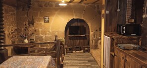 Una habitación rústica con paredes de piedra, una escalera de madera que conduce a una puerta, una mesa con mantel estampado y un microondas sobre un mueble de madera.