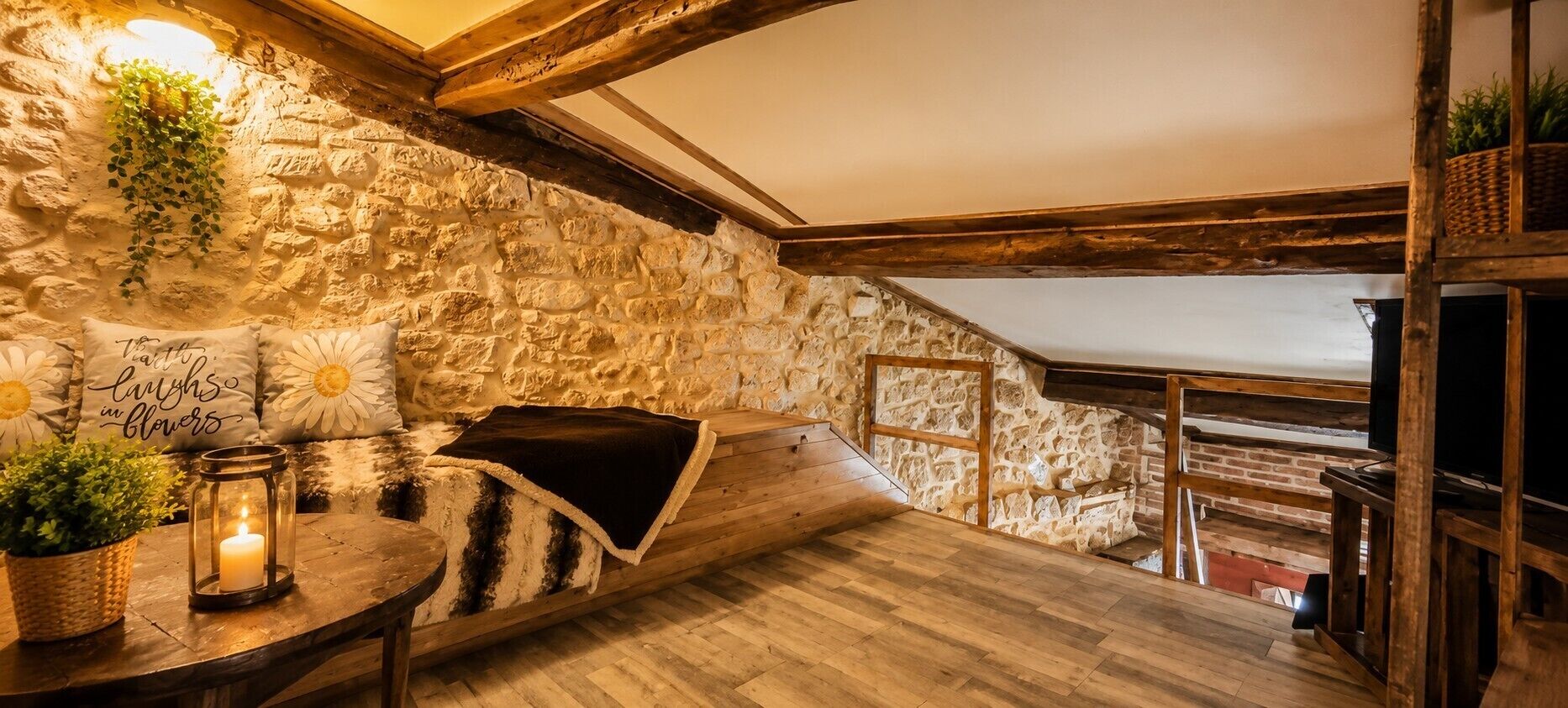 Ein gemütlicher Raum mit Steinwand, Holzbalken, einer gepolsterten Bank, einem kleinen Tisch mit Kerze und einem Blumentopf.