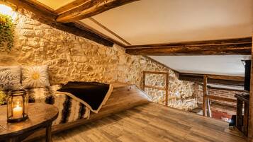 Ein gemütlicher Raum mit Steinwand, Holzbalken, einer gepolsterten Bank, einem kleinen Tisch mit Kerze und einem Blumentopf.