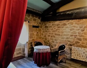 Una habitación con paredes de piedra, una cortina roja, una mesa redonda pequeña con mantel de encaje, dos sillas de mimbre y un radiador.