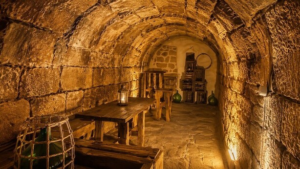 An underground stone room with a wooden table and benches, a lantern, and a stone wall.