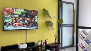 Un espace d’accueil moderne avec un mur d’accent jaune, un téléviseur à écran plat diffusant une scène de restaurant, un comptoir d’accueil agrémenté d’une plante en pot et un présentoir à magazines.