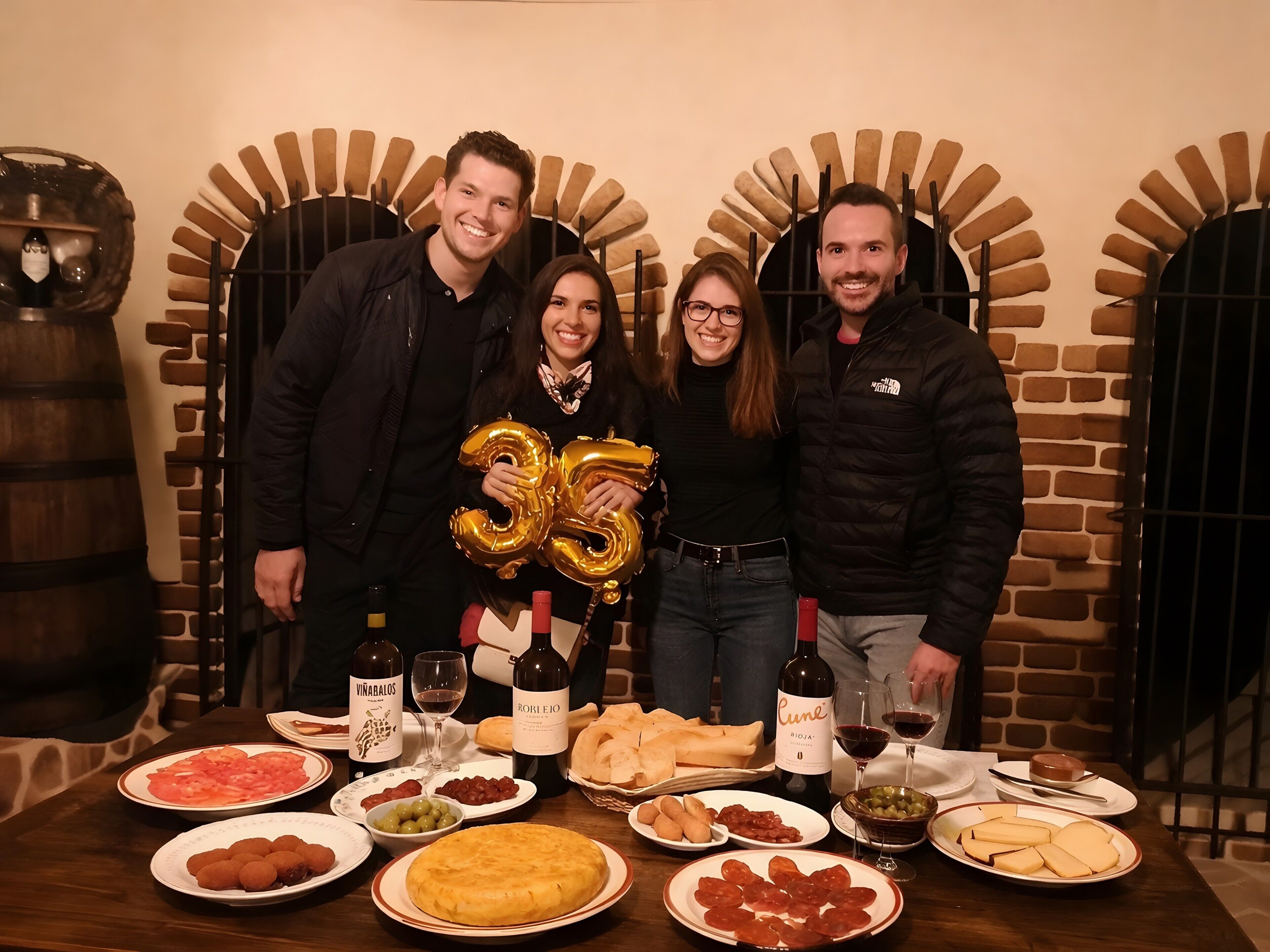 A group of people standing in front of a brick wall with arched doorways, a table with various dishes and drinks, and a bottle of wine labeled 'Cune.'