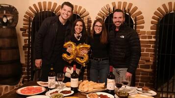 A group of people standing in front of a brick wall with arched doorways, a table with various dishes and drinks, and a bottle of wine labeled 'Cune.'