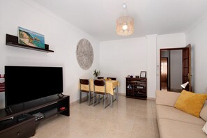 A living room with a flat-screen TV, a dining table with chairs, a sofa, a bookshelf, and a decorative wall piece.