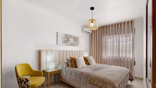 Une chambre à coucher comprenant un lit, une chaise, une table de chevet, une lampe, un luminaire au plafond et une fenêtre avec des rideaux.