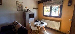 Ett litet rum med ett matbord, en vit stol, en soffa, ett element och ett fönster med gardiner.