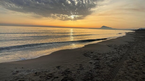Ein Strand mit Sonnenuntergang, Wellen im Meer und einem fernen Berg.