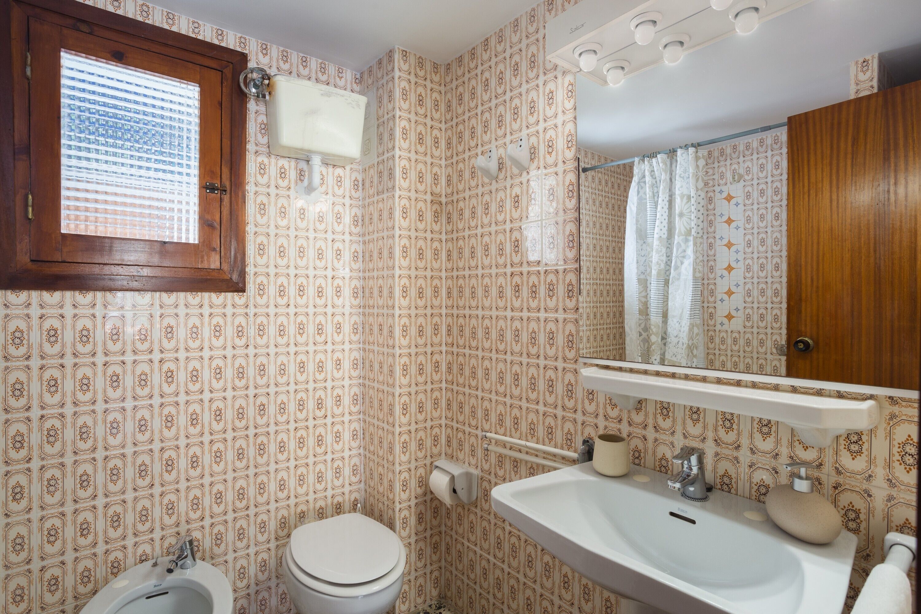 A bathroom with patterned wallpaper, a wooden-framed window, a toilet, a sink, and a mirror.